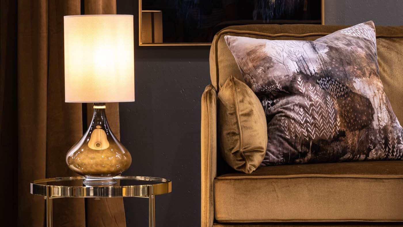 Bordlampe med hvit høy lampeskjerm stående på et gullbelagt lite sidebord og en beige fløyelssofa med en stor pute dekorert med skogsmotiv.