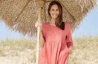 En kvinne i rosa kjole står på en strand og holder på en parasoll.