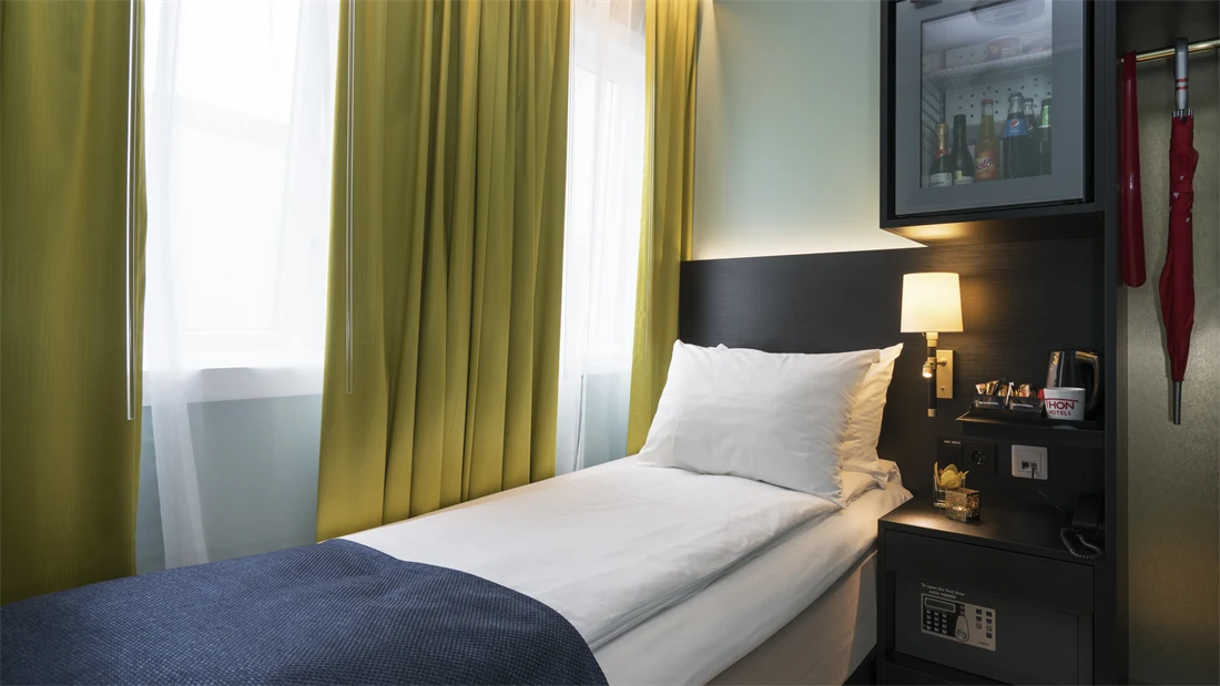 Enkeltseng med blått sengeteppe, gule gardiner og minibar på enkeltrom på Thon hotel Terminus i Oslo sentrum nær Jernbanetorget