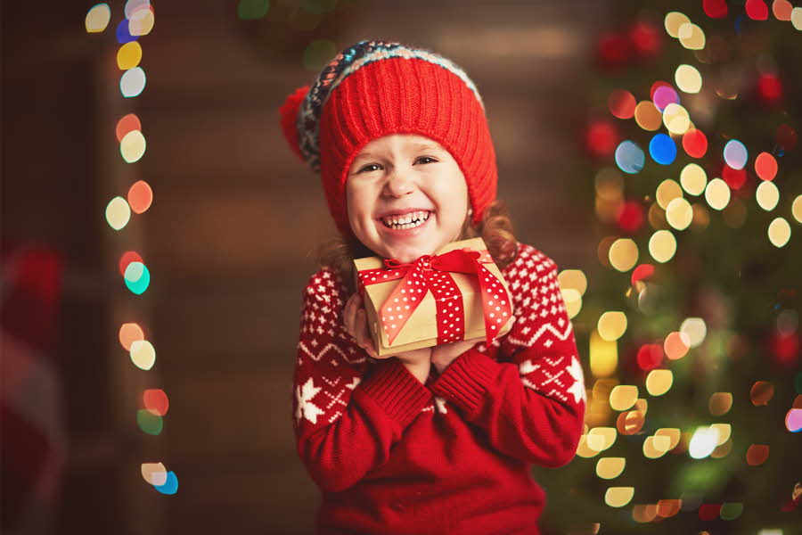 jente pyntet i rød ullgenser og rød lue, smiler og ler og holder opp julegave