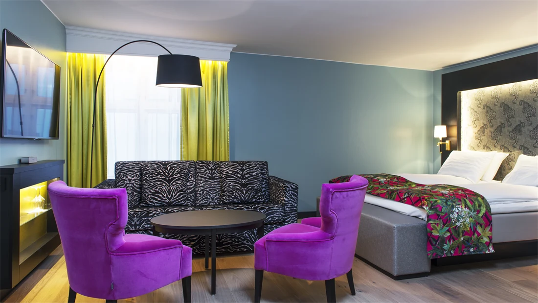 Dobbeltseng med mønstrede sengeteppe og lilla stol, zebramønstret sofa og to lilla stoler i sittegruppe, TV og gule gardiner på buisness room på Thon hotel Terminus i Oslo sentrum nær Jernbanetorget