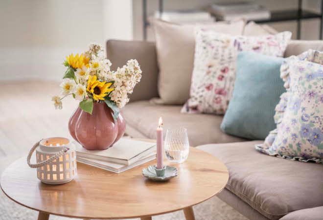 En stue med en sofa som har flere pynteputer, og et stuebord med levende lys og en vase med blomster.