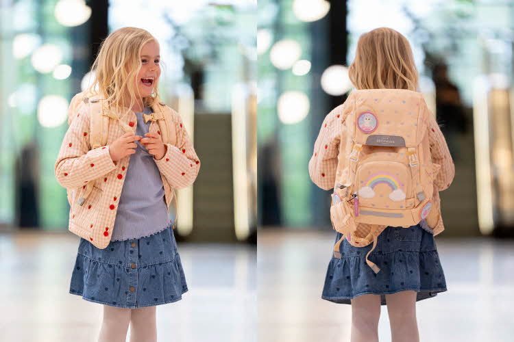 Jente med ytterjakke, olaskjørt, topp og skolesekk smiler og ser til siden. jente med ryggen mot kamera med lysegul skolesekk.