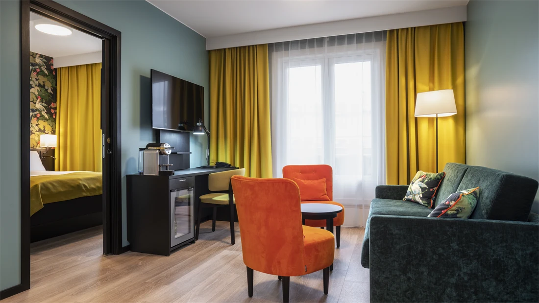 Parkettgulv, vindu fra gulv til tak, gule gardiner, turkise vegger, en grønn sofa, to oransje stoler, flatskjerm hengende på vegg, skrivebord, døråpning med innsyn til et soverom. 