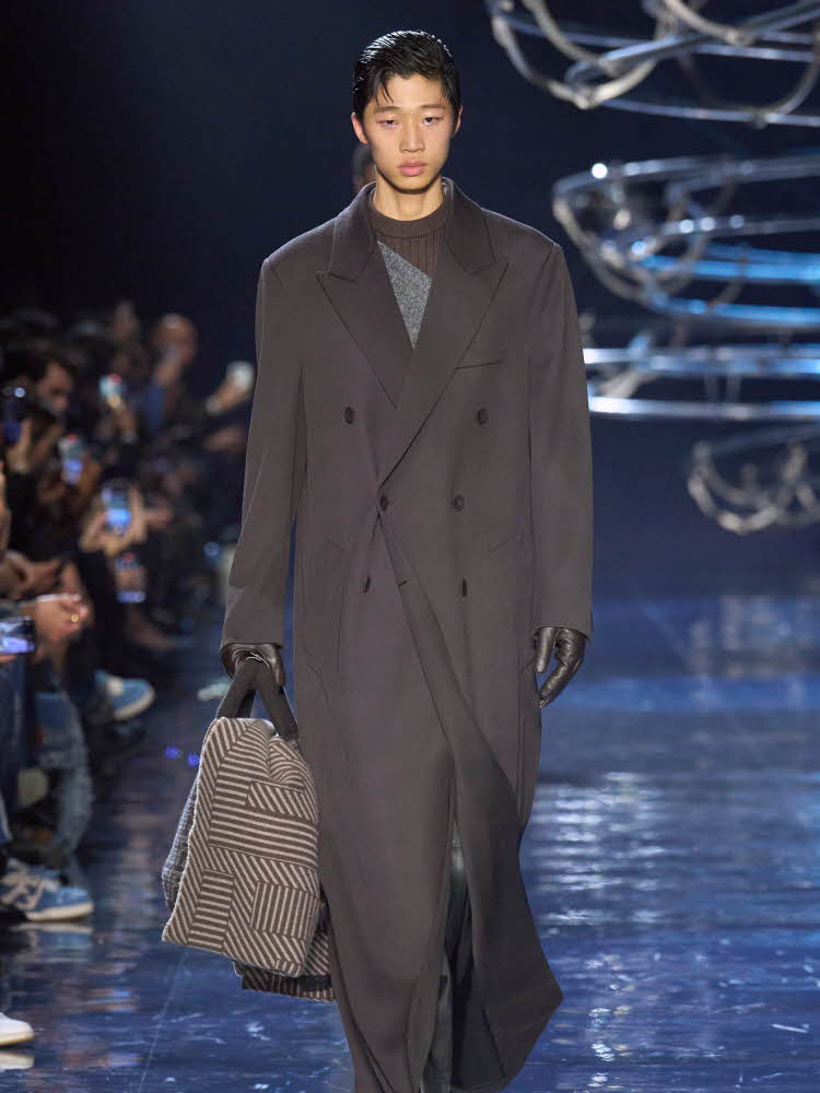 Herremodell på catwalk med mørk grå, lang kåpe og veske i hånden.