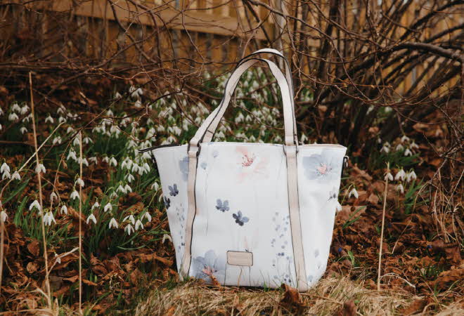 Hvit håndveske med blomsterdesign blant snøklokker og tørre blader.