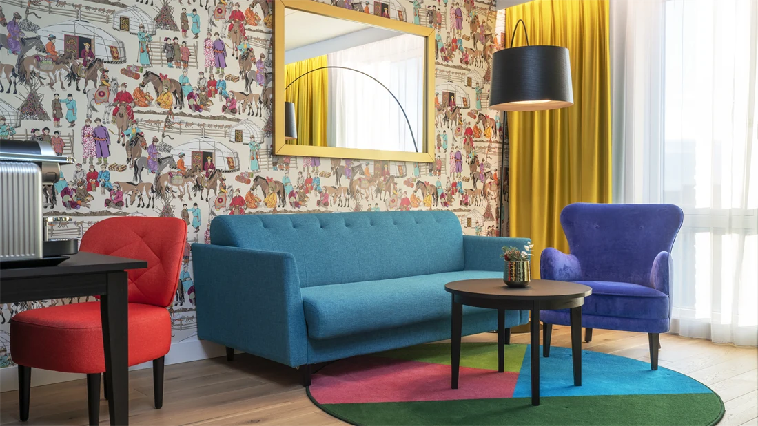 Dekorativ tapet, speil med gullramme, sittegruppe med turkis sofa, blå stol og bord på fargerikt teppe i suite  på Thon Hotel Storo