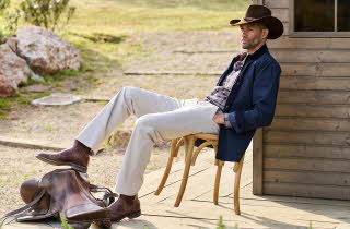 En mann i cowboy hatt sitter i en stol ute med en av føttene hans på en sal