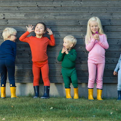 Seks barn står på en gressplen mot en husvegg kledd i ullundertøy og gummistøvler.