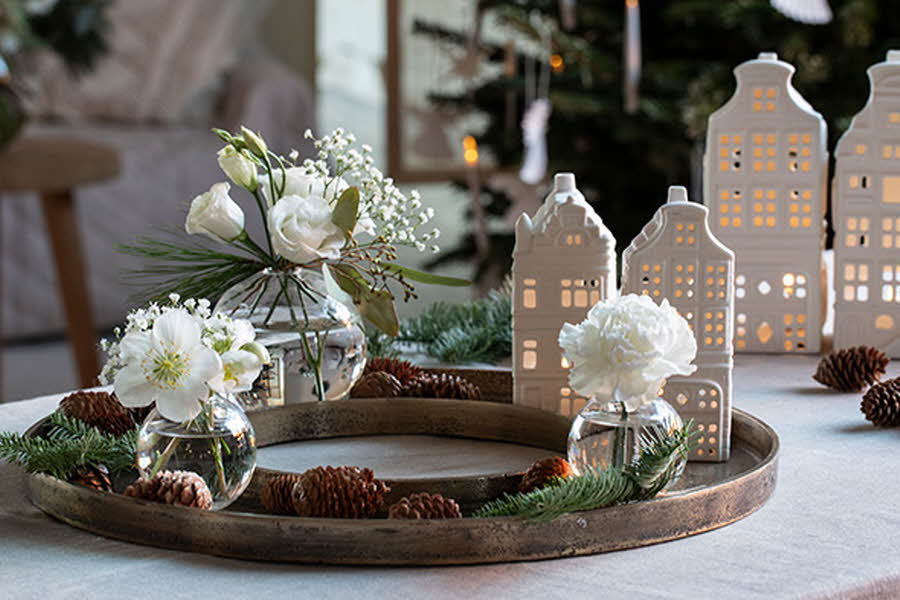 Bord pyntet med små glasskrukker med hvite blomster, grønne grener og lyshus.