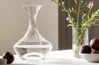 En karaffel står på et bord med en vase med blomster og en bolle med frukt.