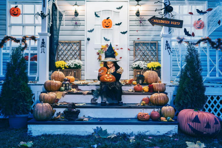 Jente, utkledd som heks til halloween, sitter på trapp i inngangsparti, omringet av gresskar og pynt.