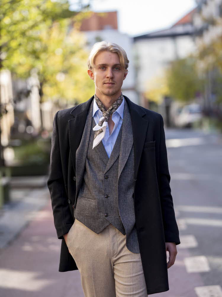 Herre i blå kåpe, grå blazer og vest, beige bukse, hvit skjorte og skjerf
