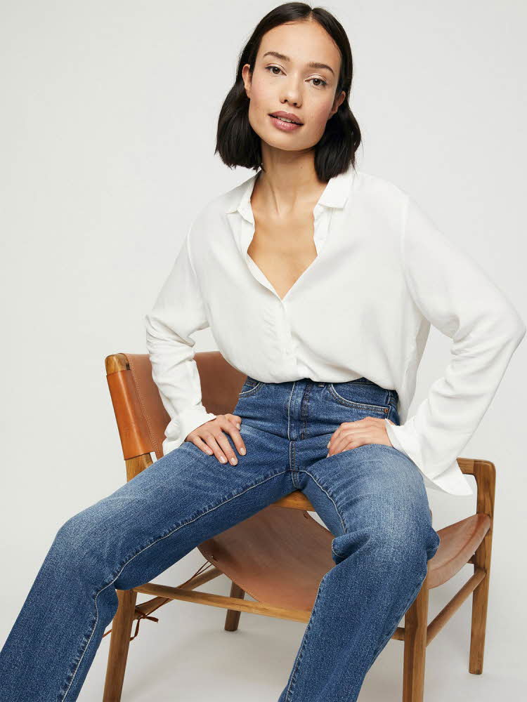 kvinnelig modell som sitter på stol iført hvit bluse og jeans
