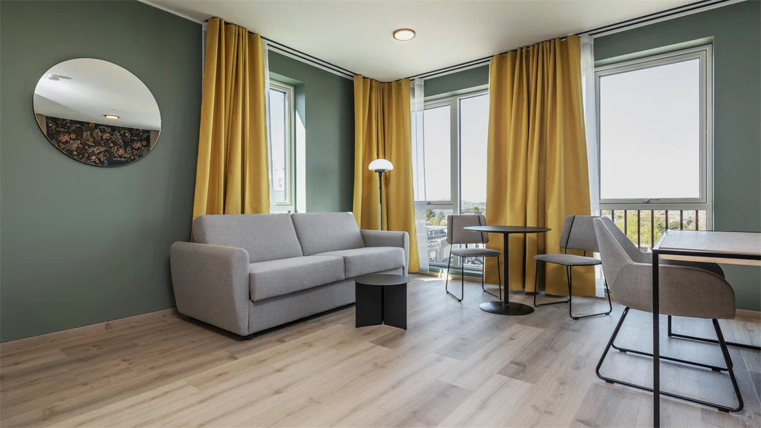 Leilighet med grå sofa, spiseplass, skrivebord med tilhørende stol, parkett, grønne vegger, store vinduer og gule gardiner.