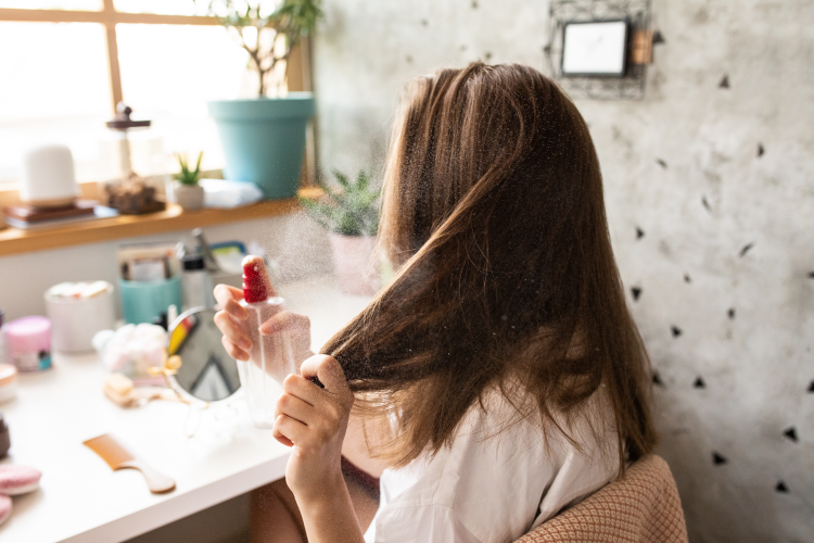  Kvinne med brunt hår sprayer håret foran et sminkebord. 