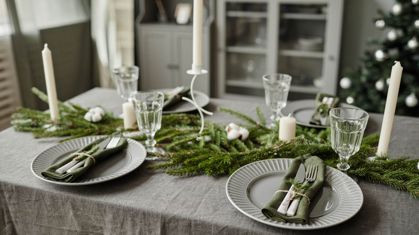 Matbord med grå linduk, pyntet med granbar og stearinlys. Fire fat, bestikk og glass. Juletre og vitrineskap i bakgrunnen.
