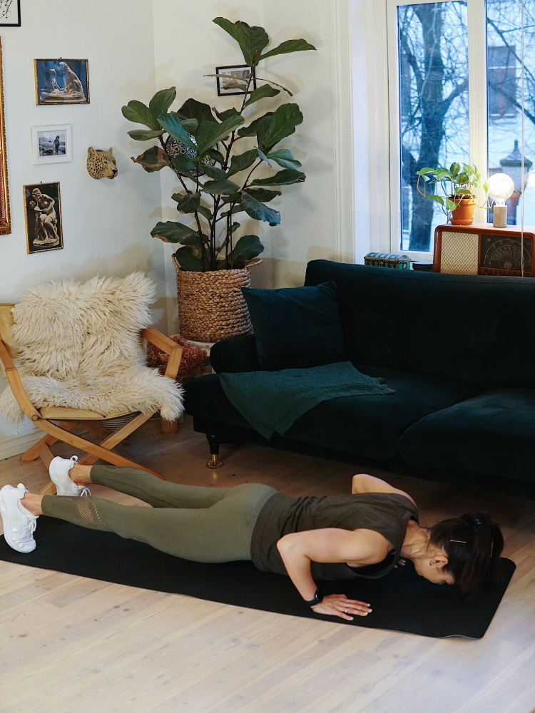 treningsekspert Anna Johansen trener på matte på gulvet, mørkegrønn sofa i bakgrunnen, lys stol med pels oppi, str grønn plante, bilder på veggen