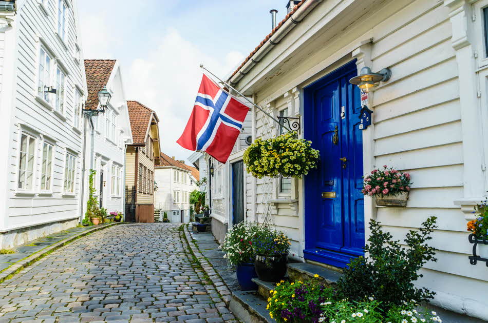 Brosteinsbelagt gate med hvite trehus, blå inngangsdør med flagg og grønne planter i forgrunn. Foto til artikkel om dekorasjoner til 17. mai.