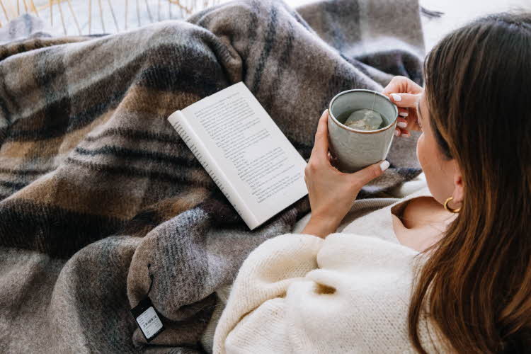Dame som ligger under pledd og leser på sofa