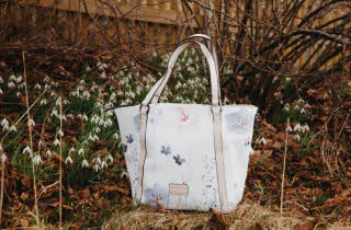 Hvit håndveske med blomsterdesign blant snøklokker og tørre blader.