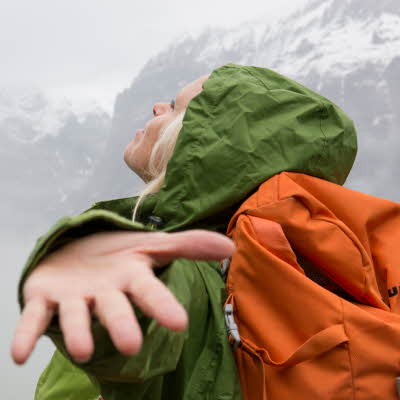 Dame i grønnregnjakke med oransje sekk holder armene ut og ser opp mot himmelen med snødekkede fjell i bakgrunnen