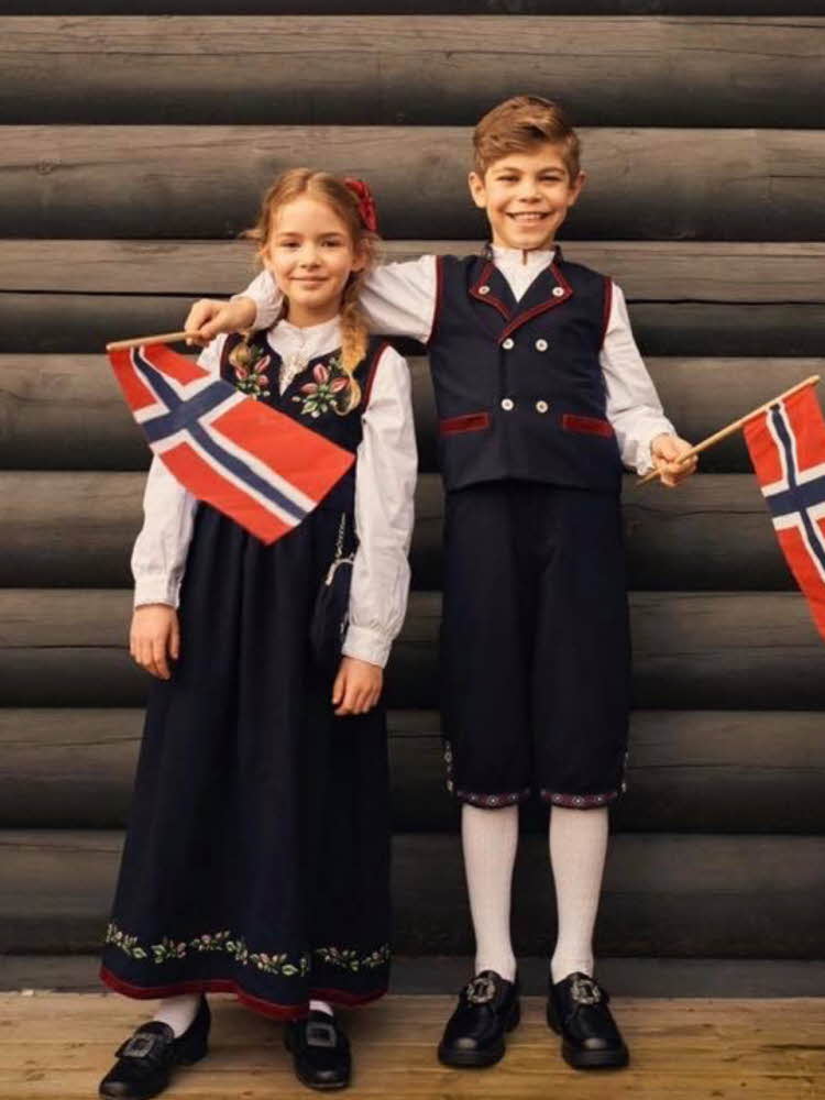 Gutt og jente med festdrakt. Gutten holder rundt jenten og har to norske flagg i hendene.