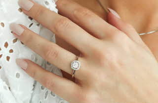 Hånd med elegant diamantring i halo-design på hvit blondebakgrunn.