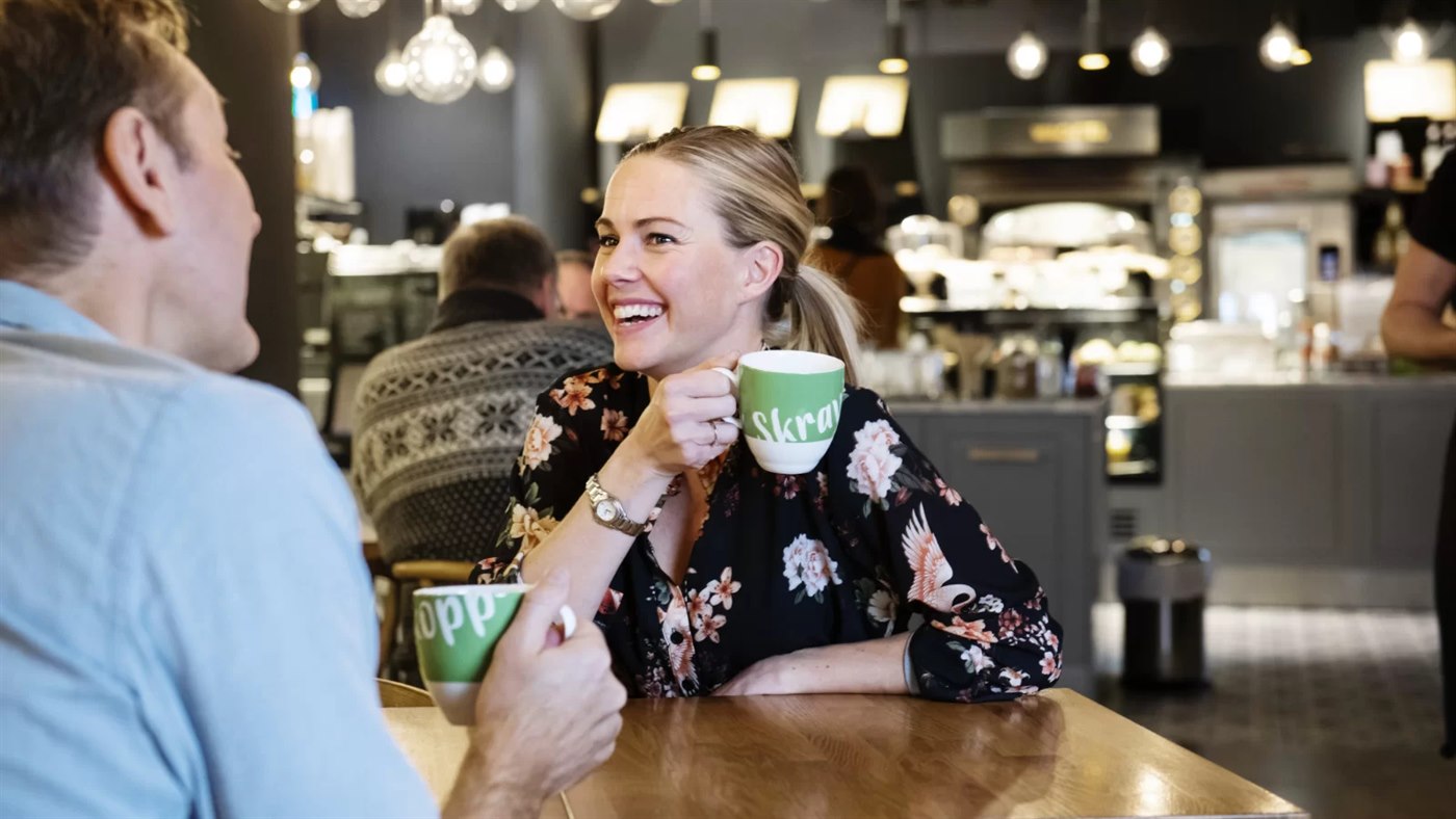 Venke Knutson sitter ved et bord og drikker kaffe fra en grønn koppe med teksten Skravlekopp på.