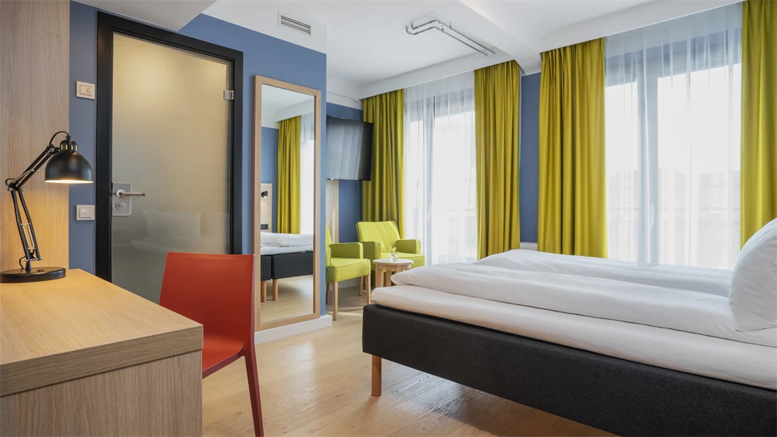 Skrivebord med rød stol, speil, blå vegger, dobbeltseng og gule gardiner i dobbeltrom på Thon Hotel Astoria