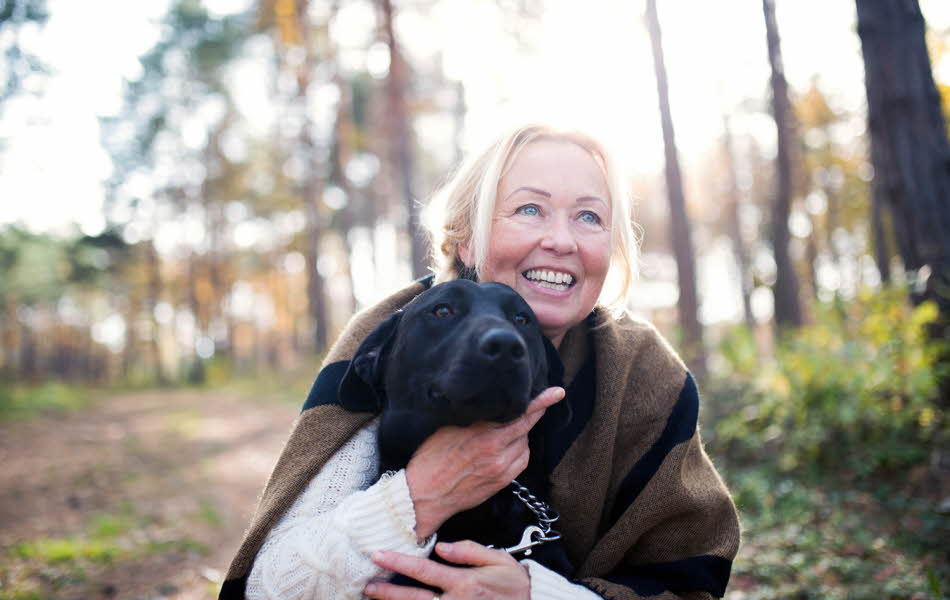 Halvportrett av smilende, middelaldrende dame som sitter på huk i skogen med en svart labrador foran seg.