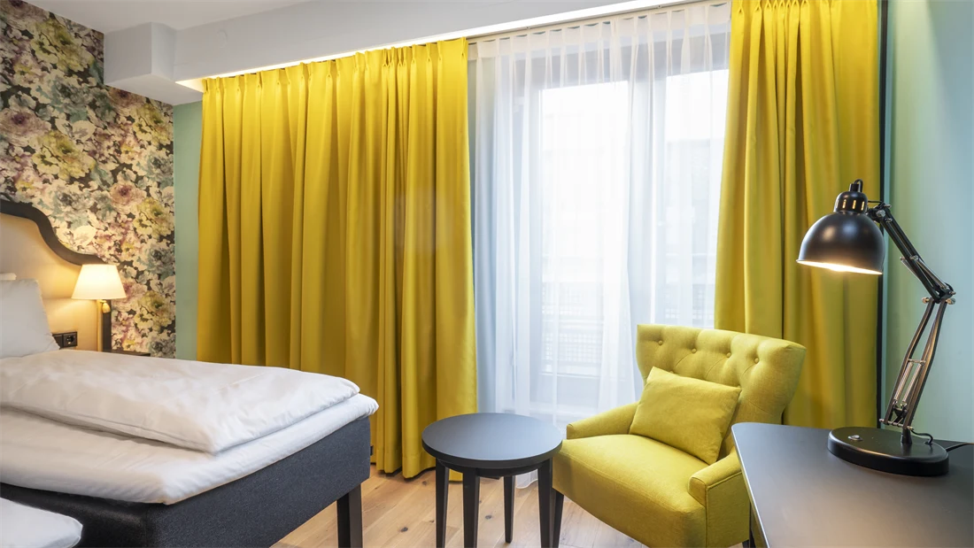 To enkeltsenger, blomstret tapet bak sengene, gul stol, arbeidspult og store vinduer med store gule gardiner på twin room på Thon Hotel Cecil i Oslo sentrum