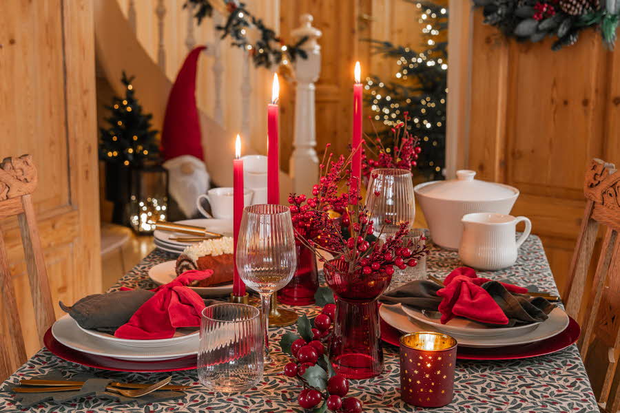 spisebord dekorert i juletema, servise, glass, stearinlys, bordtekstil med nisse og juletre i bakgrunnen