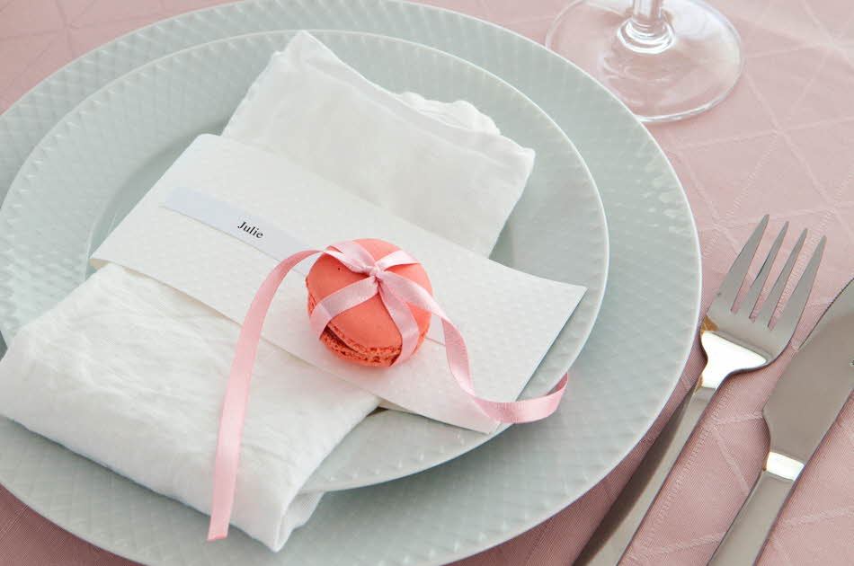 To hvite tallerker, kniv og gaffel, rosa duk og hvit serviett med rosa makron knyttet med silkebånd og en navnelapp hvor de står Julie