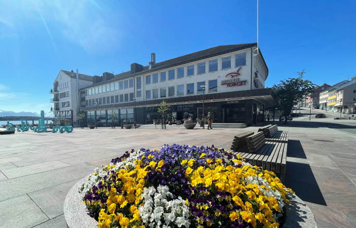 Fasadebilde av kjøpesenteret MoldeTorget. Bildet viser kjøpesenteret i bakgrunnen med sjø, blomster og kunst i forgrunnen.