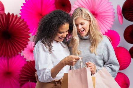 To damer med shoppingposer som ser i den ene posen og papirdekorasjon i bakgrunn