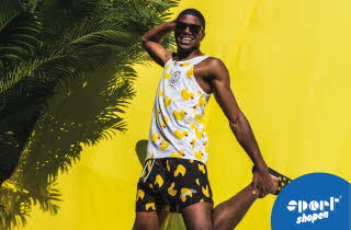 En mann i hvit singlett med badeender på og sort badeshorts med badeender på som står og poserer foran en gul vegg