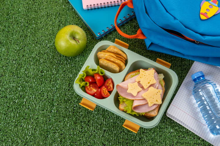 brødskiver, tomater, kjeks i en lunsjboks med granateple og vannflaske ved siden av en skolesekk og bøker