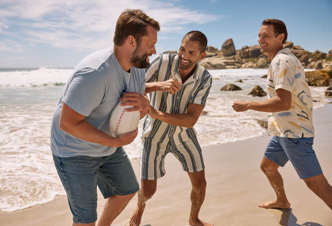 Tre menn i tskjorte og shorts spiller rugby på en strand ved vannet