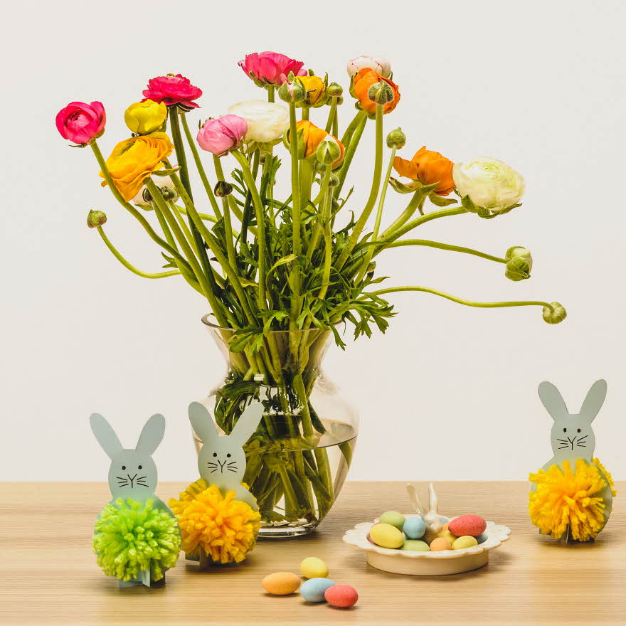 To grå kaniner med dusk laget av grønt og gult garn ved siden av en stor vase med blomster og sjokoladeegg på ett bord