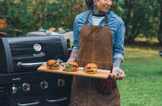 En person i brun forkle holder en treplate med tre burgere foran en Char-Broil grill.