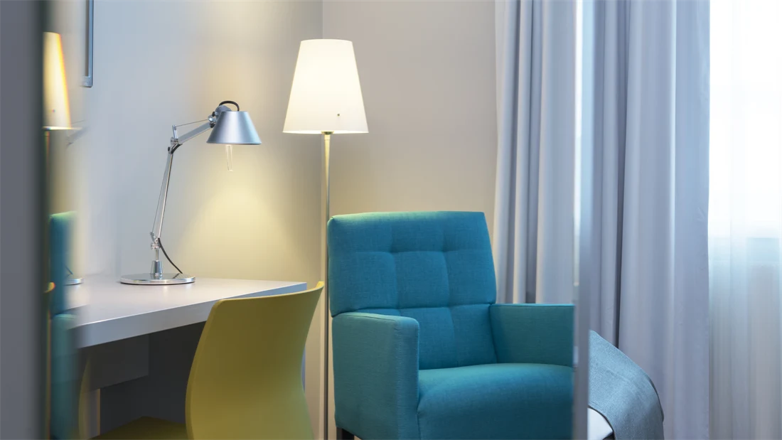 Nærbilde av Standard Single Room på Thon Hotel Trondheim. Turkis lenestol, hvitt skrivebord, gul kontorstol, leselampe, stålampe og hvite gardiner.