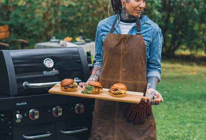 En person i brun forkle holder en treplate med tre burgere foran en Char-Broil grill.