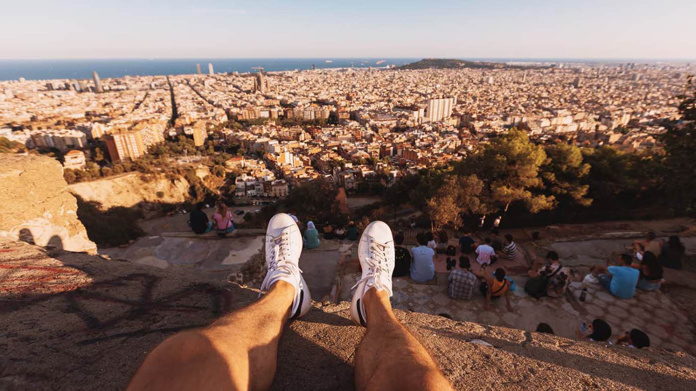 Mann sitter på en klippe. Man ser bena hans i lufta. Nedenfor er Barcelona.