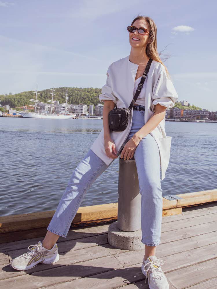 Brunette står på brygga iført lyse jeans, sort smalt belte, hvit tskjorte som er knytet og lys jakke