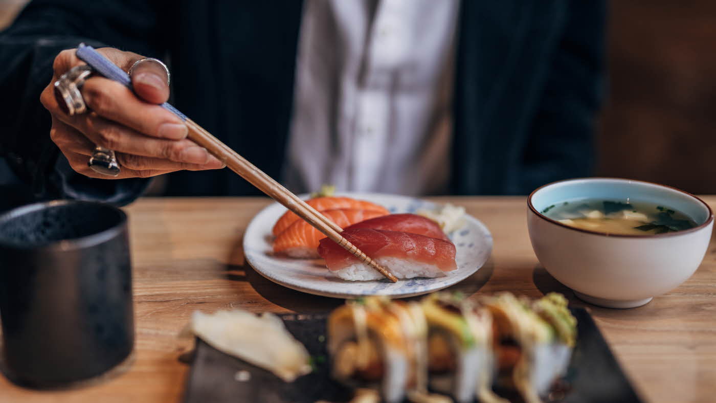 En mann holder eunfisknigiri med spisebinner på et bord med mer sushi og en bolle miso-suppe