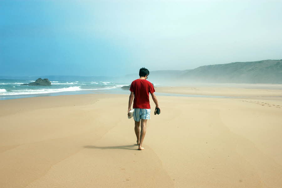 Mann på øde strand med rød t-skjorte og stripete shorts. Bølgeskvulp og sand.