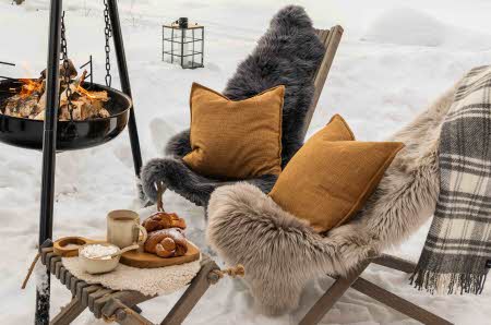 To stoler med lammeskinn og puter på sitter ute i snøen foran en bålpanne