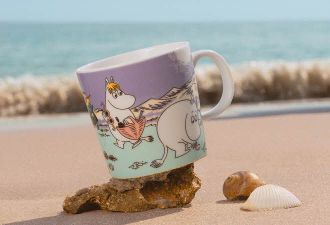 En Moomin Arabia kopp med motivet "Stranddag" står på et skjell på en strand