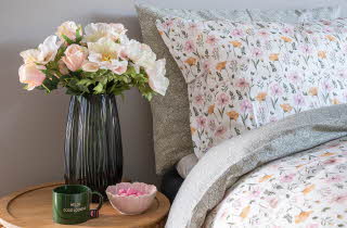 Seng med blomstrete sengetøy og nattbord med blomster og en kopp.